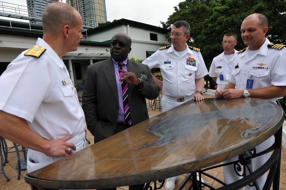 A group of five men are gathered around an ornate outdoor table, appearing to be engaged in a formal discussion or briefing. Three of the men are wearing white naval uniforms with rank insignia and ribbons, while one civilian man in a dark suit with a purple tie appears to be speaking or presenting. The setting appears to be an outdoor terrace or courtyard with buildings and trees visible in the background.