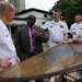 A group of five men are gathered around an ornate outdoor table, appearing to be engaged in a formal discussion or briefing. Three of the men are wearing white naval uniforms with rank insignia and ribbons, while one civilian man in a dark suit with a purple tie appears to be speaking or presenting. The setting appears to be an outdoor terrace or courtyard with buildings and trees visible in the background.