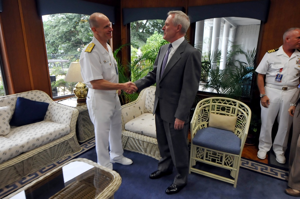 A formal meeting or greeting is taking place in what appears to be an official residence or naval facility. A naval officer in white dress uniform is shaking hands with a civilian man in a gray suit and purple tie. A second naval officer in white uniform stands in the background to the right.