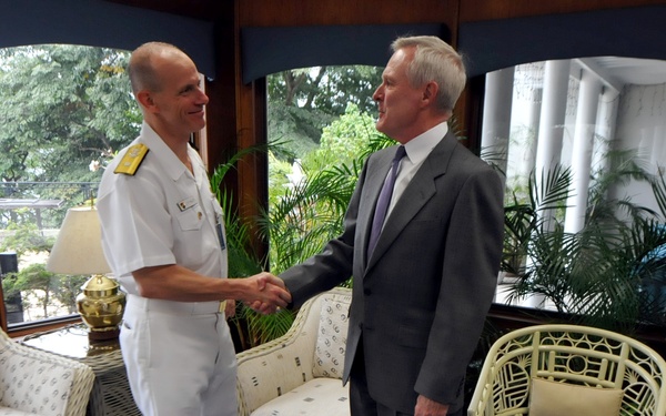 A formal meeting or greeting is taking place in what appears to be an official residence or naval facility. A naval officer in white dress uniform is shaking hands with a civilian man in a gray suit and purple tie. A second naval officer in white uniform stands in the background to the right.