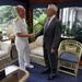 A formal meeting or greeting is taking place in what appears to be an official residence or naval facility. A naval officer in white dress uniform is shaking hands with a civilian man in a gray suit and purple tie. A second naval officer in white uniform stands in the background to the right.