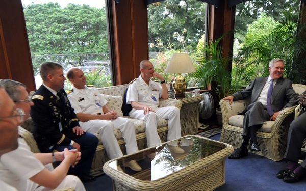 A formal meeting or discussion is taking place in a sunlit room with large windows overlooking greenery. Several individuals in U.S. military uniforms (Navy white and Army dress uniforms) are seated alongside civilians in business attire, suggesting an official diplomatic or military liaison meeting. The group is arranged in wicker/rattan furniture around a glass-topped coffee table.