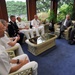 A formal meeting or discussion is taking place in a sunlit room with large windows overlooking greenery. Several individuals in U.S. military uniforms (Navy white and Army dress uniforms) are seated alongside civilians in business attire, suggesting an official diplomatic or military liaison meeting. The group is arranged in wicker/rattan furniture around a glass-topped coffee table.