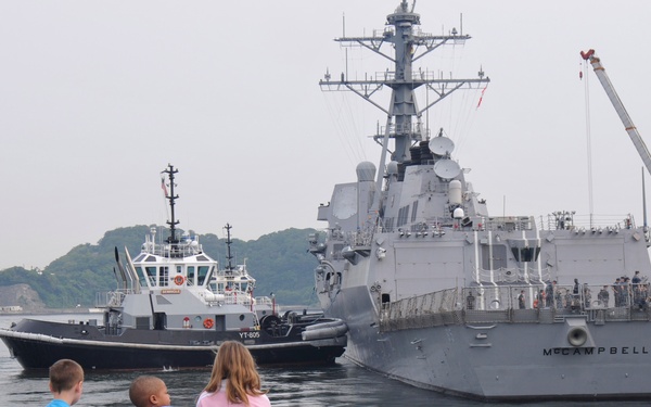 Three children sit on a stone seawall watching naval vessels in a harbor. The most prominent ship is the USS McCampbell, a U.S. Navy Arleigh Burke-class guided-missile destroyer, being assisted by a tugboat designated YT-605. The scene appears to be at a naval base, likely in Japan based on the surrounding landscape and vegetation.