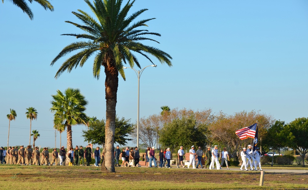 A military procession or ceremony is taking place outdoors in what appears to be a warm, coastal or southern U.S. location, evidenced by the palm trees and clear blue sky. A color guard in white Navy uniforms leads the march carrying an American flag and at least one additional flag. A large group of participants, including uniformed military personnel and civilians, follow in formation along a paved path.