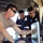 Three individuals are gathered in a confined, tarp-covered space examining a ruggedized laptop or field computer. The person on the left wearing a tan t-shirt and sunglasses appears to be demonstrating or operating the device, while two others in dark uniforms bearing what appears to be 'Policía Nacional' patches observe closely. The setting suggests a field or operational environment, possibly a military or law enforcement training or coordination scenario.