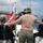 A group of people on a dive boat preparing for a scuba diving excursion on calm coastal waters. A central figure wearing a tan t-shirt with 'Construction' text and a camouflage hat is assisting divers with their equipment. A red-and-white diver-down flag is mounted on a pole at the stern of the boat.