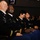 Four U.S. Army personnel in dress black uniforms are seated in an auditorium-style venue, laughing and smiling while looking upward, likely watching a presentation or ceremony. Name tags visible on their uniforms read Brining, Culbreth, Gardner, and Gonzales. Additional audience members are visible in the background.