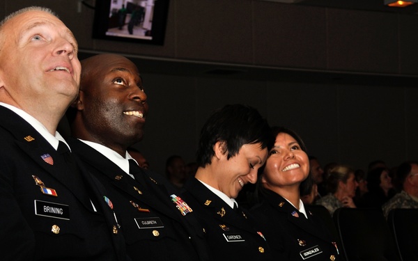Four U.S. Army personnel in dress black uniforms are seated in an auditorium-style venue, laughing and smiling while looking upward, likely watching a presentation or ceremony. Name tags visible on their uniforms read Brining, Culbreth, Gardner, and Gonzales. Additional audience members are visible in the background.