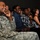 A group of people seated in an auditorium or theater are laughing and smiling, appearing to enjoy a performance or presentation. The foreground features a U.S. Army soldier in ACU (Army Combat Uniform) with an American flag patch visible on the left sleeve. The audience includes both military personnel and civilians, including children, suggesting a family-friendly military event.