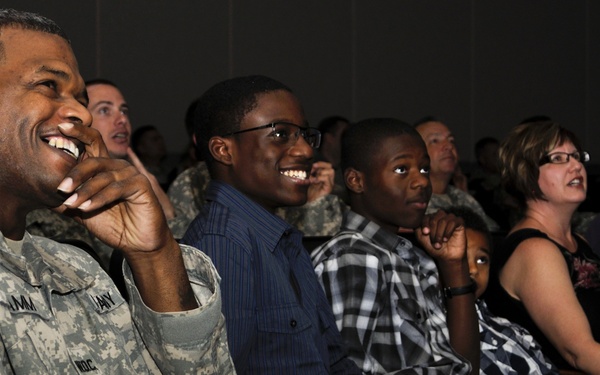 A group of people seated in an auditorium or theater are laughing and smiling, appearing to enjoy a performance or presentation. The foreground features a U.S. Army soldier in ACU (Army Combat Uniform) with an American flag patch visible on the left sleeve. The audience includes both military personnel and civilians, including children, suggesting a family-friendly military event.