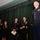 A U.S. Army military ceremony is taking place on a stage with green curtains in the background. Five uniformed Army personnel are present, with one officer in dress blues standing to the right appearing to be the honoree, while four others in Army Service Uniforms (ASUs) applaud or stand at attention. The setting suggests a promotion, retirement, or award ceremony.