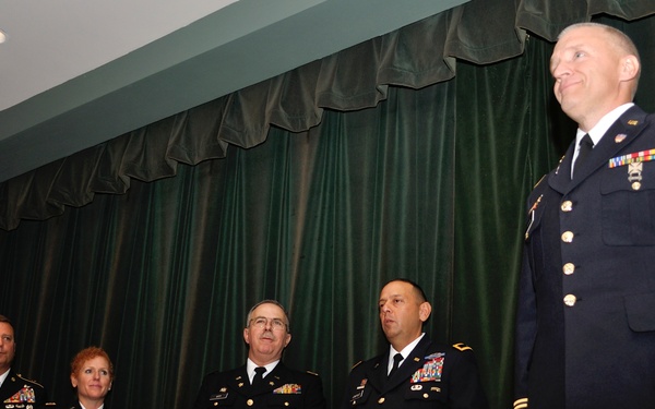 A U.S. Army military ceremony is taking place on a stage with green curtains in the background. Five uniformed Army personnel are present, with one officer in dress blues standing to the right appearing to be the honoree, while four others in Army Service Uniforms (ASUs) applaud or stand at attention. The setting suggests a promotion, retirement, or award ceremony.