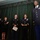A formal U.S. Army military ceremony taking place on a stage with green curtain backdrop. Five uniformed Army personnel are standing on stage, with one individual prominently positioned at the front right, appearing to be the focus of recognition. Several personnel are holding documents, suggesting an awards or promotion ceremony.