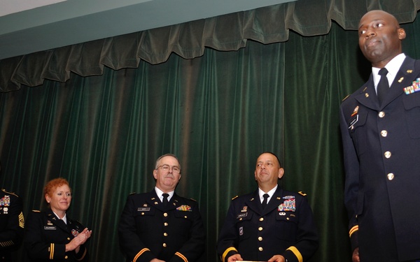 A formal U.S. Army military ceremony taking place on a stage with green curtain backdrop. Five uniformed Army personnel are standing on stage, with one individual prominently positioned at the front right, appearing to be the focus of recognition. Several personnel are holding documents, suggesting an awards or promotion ceremony.