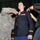 A military promotion ceremony is taking place, with a female U.S. Army officer in dress blues having new rank insignia pinned onto her shoulders simultaneously by two individuals — one in ACU (Army Combat Uniform) on her left and a civilian on her right. The officer stands at attention with a composed expression, looking upward, as is traditional during a pinning ceremony. Additional uniformed personnel are visible in the background.