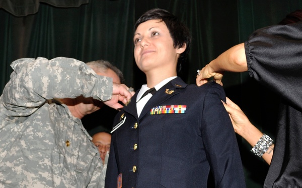 A military promotion ceremony is taking place, with a female U.S. Army officer in dress blues having new rank insignia pinned onto her shoulders simultaneously by two individuals — one in ACU (Army Combat Uniform) on her left and a civilian on her right. The officer stands at attention with a composed expression, looking upward, as is traditional during a pinning ceremony. Additional uniformed personnel are visible in the background.