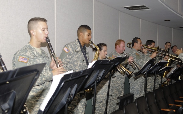 A group of U.S. Army soldiers in ACU (Army Combat Uniform) camouflage are performing as a military band in what appears to be an auditorium or briefing room. The musicians are arranged in a line with music stands in front of them, playing various wind instruments while a conductor on the right directs them. The setting suggests a rehearsal or live performance at a military facility.