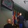 A military ceremony or event is taking place in an indoor venue. Two uniformed military personnel are visible from behind — one standing on a raised platform holding a microphone near a display of flags, and another standing at attention in the foreground. A wall-mounted television screen displays an image of a group of people, likely soldiers or civilians, in green shirts.
