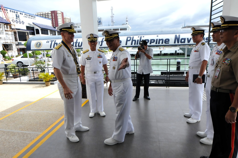 A group of naval officers in white dress uniforms are gathered at what appears to be a Philippine Navy facility, visible from the text 'The Future of the Philippine Navy' on a vessel or banner in the background. One officer appears to be gesturing and briefing or directing another senior officer, suggesting an official visit or inspection. A photographer is capturing the event in the background.