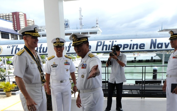 A group of naval officers in white dress uniforms are gathered at what appears to be a Philippine Navy facility, visible from the text 'The Future of the Philippine Navy' on a vessel or banner in the background. One officer appears to be gesturing and briefing or directing another senior officer, suggesting an official visit or inspection. A photographer is capturing the event in the background.