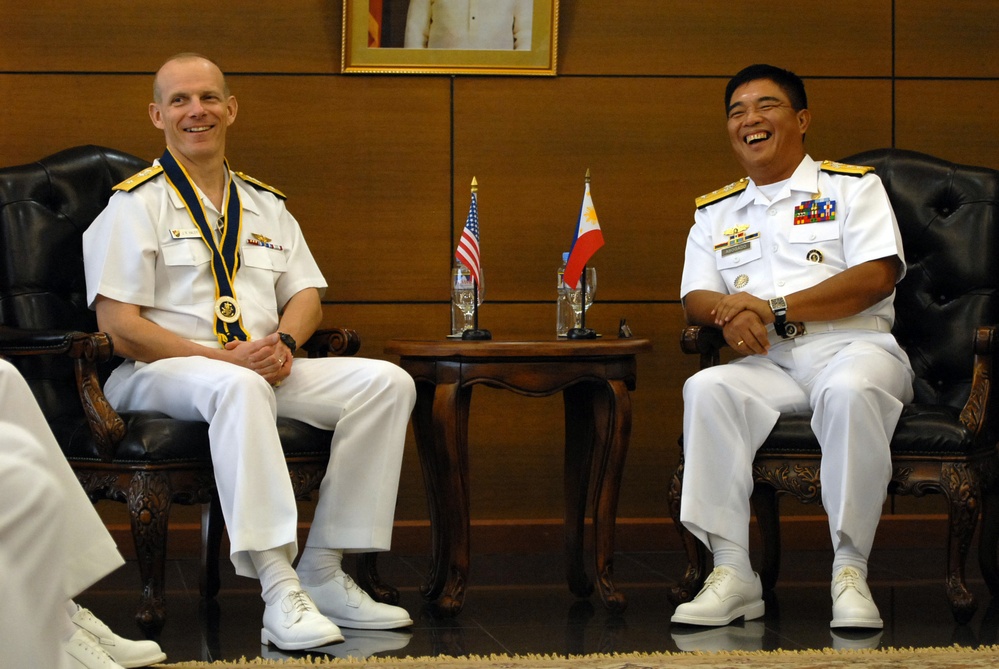 Two naval officers in white dress uniforms are seated in ornate leather chairs, laughing and smiling during what appears to be a formal bilateral meeting. Between them is a small table displaying miniature American and Philippine flags, suggesting a US-Philippines military diplomatic engagement. A framed portrait hangs on the wood-paneled wall behind them.