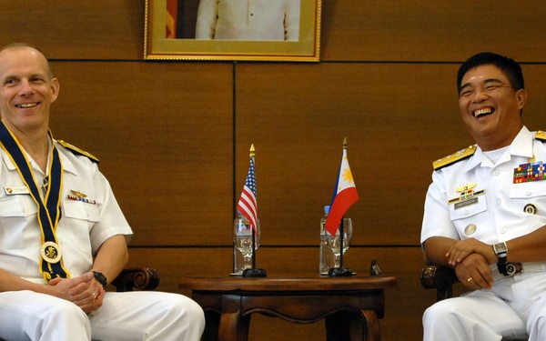 Two naval officers in white dress uniforms are seated in ornate leather chairs, laughing and smiling during what appears to be a formal bilateral meeting. Between them is a small table displaying miniature American and Philippine flags, suggesting a US-Philippines military diplomatic engagement. A framed portrait hangs on the wood-paneled wall behind them.