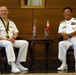 Two naval officers in white dress uniforms are seated in ornate leather chairs, laughing and smiling during what appears to be a formal bilateral meeting. Between them is a small table displaying miniature American and Philippine flags, suggesting a US-Philippines military diplomatic engagement. A framed portrait hangs on the wood-paneled wall behind them.