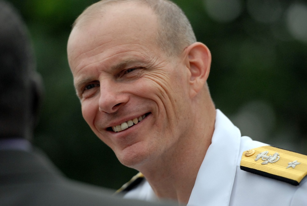 A smiling middle-aged man wearing a white military dress uniform with gold and silver shoulder epaulettes is photographed in close-up. The uniform insignia suggests a senior officer rank, likely naval based on the style of the epaulettes. The background is blurred green foliage, indicating an outdoor setting.