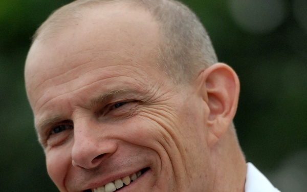 A smiling middle-aged man wearing a white military dress uniform with gold and silver shoulder epaulettes is photographed in close-up. The uniform insignia suggests a senior officer rank, likely naval based on the style of the epaulettes. The background is blurred green foliage, indicating an outdoor setting.