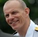 A smiling middle-aged man wearing a white military dress uniform with gold and silver shoulder epaulettes is photographed in close-up. The uniform insignia suggests a senior officer rank, likely naval based on the style of the epaulettes. The background is blurred green foliage, indicating an outdoor setting.