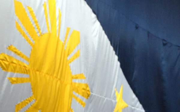 A man in a gray suit is speaking at a microphone podium in front of a large Philippine flag displayed as a backdrop. The flag features the distinctive sun with eight rays, three stars, and the blue, red, and white tricolor design. The setting appears to be a formal or official event venue.