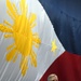 A man in a gray suit is speaking at a microphone podium in front of a large Philippine flag displayed as a backdrop. The flag features the distinctive sun with eight rays, three stars, and the blue, red, and white tricolor design. The setting appears to be a formal or official event venue.