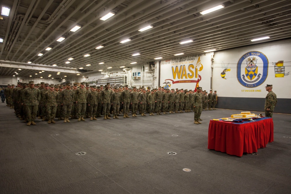 A large military formation of U.S. Marines is assembled in the hangar bay of USS Wasp (LHD-1), an amphibious assault ship. The personnel are standing at attention in rows facing a ceremonial table draped in red cloth, suggesting a formal military ceremony or celebration. The ship's insignia, name, and 'Stinger One' nickname are prominently displayed on the hangar bay wall.