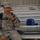 A male soldier in US Army ACU (Army Combat Uniform) sits on metal bleachers, drinking from a hydration system tube. He is wearing a patrol cap and has a backpack on, with unit patches visible on his left sleeve including a US flag patch. Various personal items including a blue lunch bag, a drink cup, and other gear are visible on the bleachers nearby.