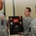 Three U.S. Army soldiers in ACU (Army Combat Uniform) digital camouflage are participating in a military retirement or award ceremony. A shadow box plaque bearing the name 'CW5 Michael Hayduk' with dates '1988-2012' is being presented, containing military patches, ribbons, and a guidon flag. The soldier on the left wears a 10th Mountain Division patch, indicating unit affiliation.