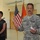 A U.S. Army soldier, identified by name tape as 'HAIDUK' (or similar spelling), is speaking at what appears to be an official military ceremony or briefing inside a large indoor facility. He is holding a small piece of paper and gesturing while addressing an audience. A civilian woman in a dark floral blouse stands behind him near a podium, with an American flag and an orange military unit flag visible in the background.