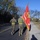 Two U.S. Marines in camouflage utility uniforms and yellow safety belts are walking along a road, with one carrying a red United States Marine Corps organizational flag with gold fringe. The scene appears to take place on a military installation or base, evidenced by the brick buildings visible in the background. The setting is a clear autumn day with bare and partially leafed trees lining the road.