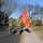 Two United States Marines in combat utility uniforms walk along a road and sidewalk carrying a red United States Marine Corps guidon or organizational flag. They are wearing olive drab long-sleeve shirts with MARPAT camouflage trousers and yellow safety belts, suggesting a physical training or ceremonial run. The setting appears to be a military installation or campus during autumn.