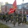 A large formation of United States Marines in PT (physical training) gear is conducting a unit run along a sidewalk on what appears to be a military installation. A Marine in the front carries a red United States Marine Corps guidon flag. The setting features classic red brick colonial-style buildings typical of established East Coast military bases.