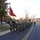 A formation of United States Marines in camouflage uniforms and yellow safety belts conducts a unit run along a paved road on what appears to be a military installation. A Marine at the front carries a red USMC guidon flag while leading the formation. The setting appears to be autumn based on the foliage, with military and civilian buildings visible in the background.