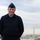 A male military personnel member in US Air Force dress uniform stands outdoors smiling, with the Washington DC skyline visible in the background including the Washington Monument. The photo appears to be a formal or semi-formal portrait taken from an elevated vantage point overlooking the Potomac River area. The overcast sky and autumn foliage suggest the photo was taken in fall.