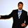 A man in a dark suit and blue striped tie is wearing what appears to be a Medal of Honor on a blue ribbon around his neck. He is gesturing with his left arm extended outward with a thumbs-up or pointing gesture, suggesting he is giving a speech or presentation. The background is a plain light-colored wall or projection screen.