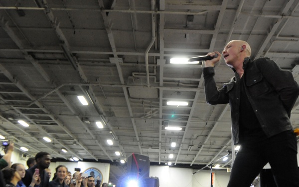 A bald male performer dressed entirely in black is singing into a handheld microphone on a raised stage, performing for a large crowd in what appears to be the hangar bay of a military vessel. The audience includes individuals in military uniforms as well as civilians, many of whom are photographing or filming the performance. A grand piano is visible in the background on the right side of the stage.