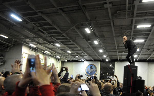 A live musical performance is taking place inside what appears to be a naval vessel's hangar bay or interior deck. A performer stands elevated on top of a speaker stack, holding a microphone, while a large crowd of audience members — many in U.S. Navy uniforms — photograph and record the event with phones and cameras. A guitarist is visible on the stage to the right, and a military insignia/emblem is mounted on the wall in the background.