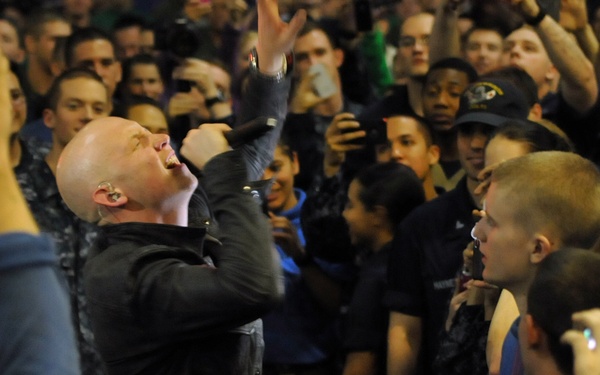 A bald male performer is singing into a microphone while surrounded by a large crowd at what appears to be an indoor concert or live performance event. He is wearing a black jacket and has one arm raised dramatically while tilting his head back. The audience is densely packed around him, with many people recording the performance on smartphones and mobile devices.
