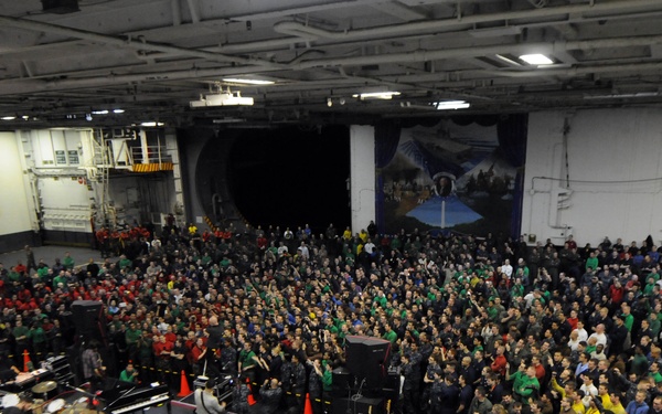 A large indoor concert or entertainment event is taking place inside what appears to be a U.S. Navy aircraft carrier hangar bay, identifiable by the 'CTF-70' insignia on the upper left wall. Hundreds of military personnel in various colored uniforms are gathered as an audience facing a stage with musical equipment including drums, speakers, and instruments. The event appears to be a morale-boosting USO-style performance for the ship's crew.
