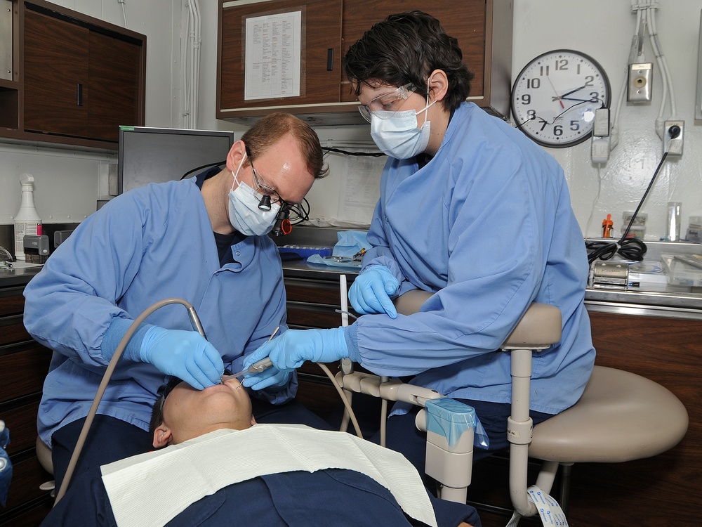 Two dental professionals in blue scrubs, surgical masks, and gloves are performing a dental procedure on a reclined patient. The setting is a clinical dental operatory with standard equipment visible. One practitioner appears to be the primary dentist using magnification loupes, while the other serves as a dental assistant.