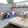 A military personnel in Marine Corps camouflage uniform stands on a wooden platform/dock addressing a group of approximately 20 civilians. The setting appears to be a training or demonstration facility with concrete block walls and a wooden overhead structure. The group appears to be on an organized tour or briefing at what looks like a military installation.