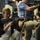 A woman in a black cardigan and white top is handling an AR-15 style rifle, appearing to examine or aim it in what looks like an indoor demonstration or media event. Several other individuals are visible in the background, some also handling similar firearms or operating camera equipment. The setting appears to be an organized event, possibly a press demonstration or firearms exhibition.